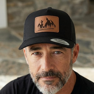 Puede incluir: Gorra de camionero negra con un parche de cuero marrón con un diseño de montaña y bosque. La gorra tiene una parte trasera de malla y una visera curva. El parche es rectangular y está cosido en la parte delantera.