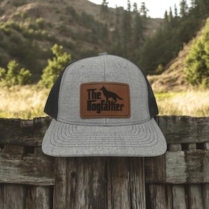 May include: A gray and black trucker hat with a brown leather patch that says "The Dogfather" with a silhouette of a German Shepherd dog.