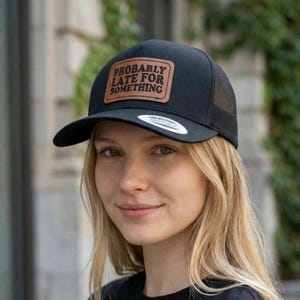 May include: Black trucker hat with a curved brim and mesh back. The hat features a brown leather patch with the words "PROBABLY LATE FOR SOMETHING" in a serif font. The hat is a Yupoong brand.