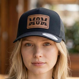 Peut inclure: Casquette de camionneur noire avec un patch en cuir marron portant l'inscription "MOM" et "Mila Rose" au-dessus. La casquette a une visière incurvée et un dos en filet.