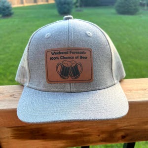 May include: A grey and black trucker hat with a brown leather patch that reads "Weekend Forecast: 100% Chance of Beer" with a graphic of two beer mugs clinking.