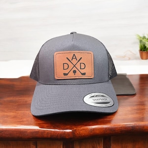 Op de afbeelding: Een grijze en zwarte trucker-pet met een bruin leren patch waarop "A D X D" staat met gekruiste golfclubs en een golfbal. De pet heeft een zwart mesh-achterpaneel en een "The Authentic Snapback"-label.
