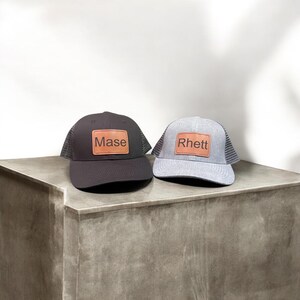 Puede incluir: Dos gorras de camionero negras y grises con parches de cuero. El parche de la gorra negra dice "Mase" y el parche de la gorra gris dice "Rhett".