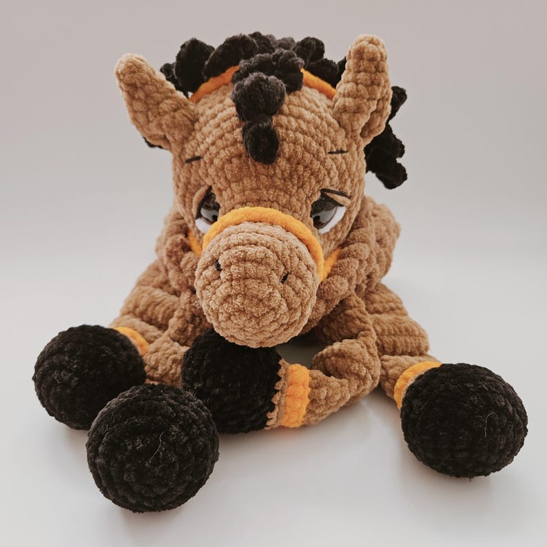 Crochet brown horse pajama bag plush toy, lying down and looking straight ahead, black mane and orange bridle details