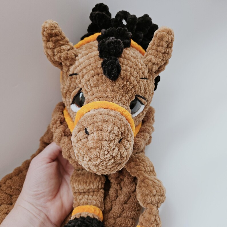 Handmade crochet horse pajama holder in brown, soft plush toy with black hooves and orange bridle, shown held in a human hand for size reference, close face photo