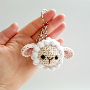 May include: A white crocheted lamb keyring with black eyes and a pink nose. The lamb is attached to a silver key ring.