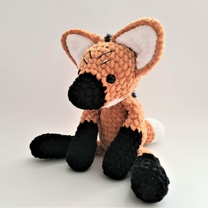 May include: A crocheted stuffed animal resembling a brown and black maned wolf with white ears and a black nose. The toy is sitting on a white surface.