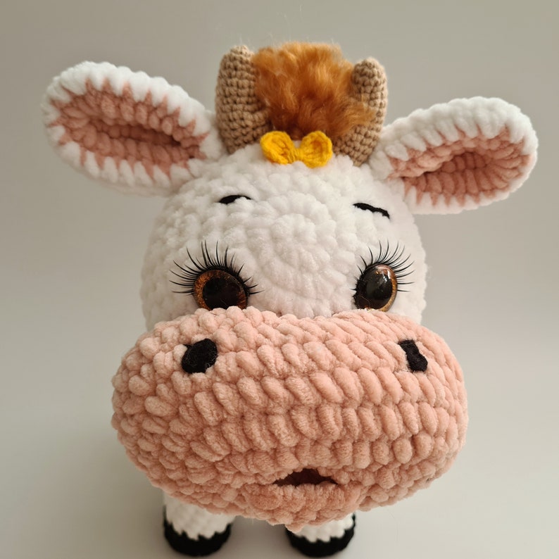 May include: A crocheted plush toy cow with white fur, brown and pink accents, and large brown eyes. The cow has a yellow bow on its head and a pink nose.