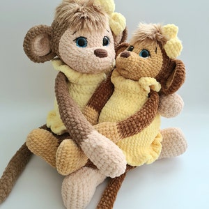 May include: Two crocheted monkey plush toys, one brown and one yellow, hugging each other. The larger monkey is brown with yellow yarn accents and the smaller monkey is yellow with brown yarn accents. Both monkeys have black eyes and brown yarn noses.
