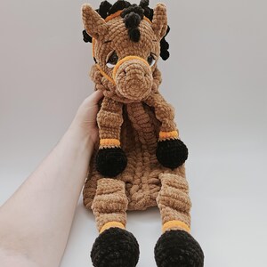 Handmade crochet horse pajama holder in brown, soft plush toy with black hooves and orange bridle, shown held in a human hand for size reference