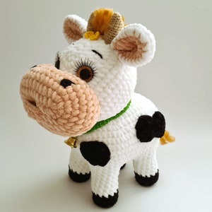 May include: A crocheted plush toy cow with white fur, black spots, and a green collar with a bell. The cow has a brown nose, brown eyes, and a yellow tuft of hair on its head.