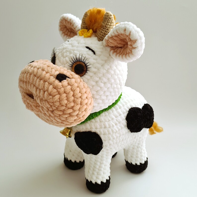 May include: A crocheted plush toy cow with white fur, black spots, and a green collar with a bell. The cow has a brown nose, brown eyes, and a yellow tuft of hair on its head.