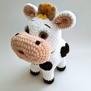 May include: A white crocheted cow plush toy with black spots, brown ears, and a brown and yellow flower on its head. The cow has a pink nose and black eyes.