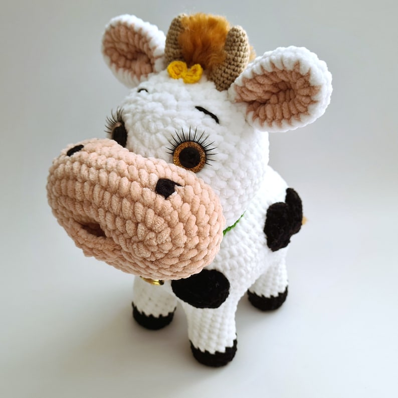 May include: A white crocheted cow plush toy with black spots, brown ears, and a brown and yellow flower on its head. The cow has a pink nose and black eyes.