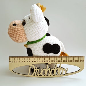 May include: A crocheted white cow with black spots and a green collar. The cow is sitting on a wooden ruler with the word "Divohobby" printed on it.