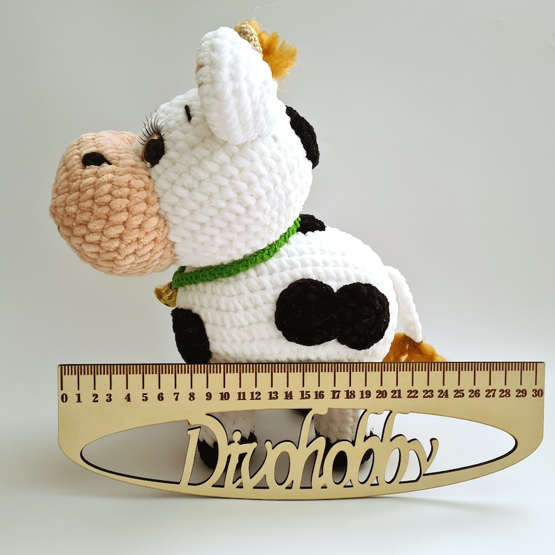 May include: A crocheted white cow with black spots and a green collar. The cow is sitting on a wooden ruler with the word "Divohobby" printed on it.