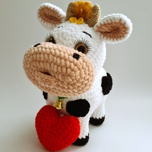 May include: A white crocheted cow plush toy with black spots, holding a red crocheted heart. The cow has a brown nose and black eyes with gold glitter. The cow has a yellow bow on its head.
