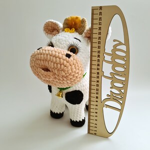 May include: A white crocheted cow plush toy with black spots, a pink nose, and a green bell around its neck. The toy is approximately 18 inches tall and is standing next to a wooden ruler.