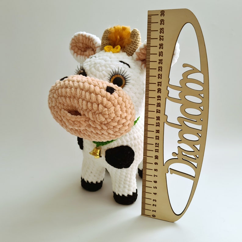 May include: A white crocheted cow plush toy with black spots, a pink nose, and a green bell around its neck. The toy is approximately 18 inches tall and is standing next to a wooden ruler.