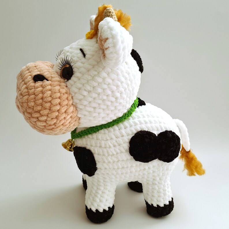 May include: A white crocheted cow plush toy with black spots, a green collar, and a gold bell. The cow has a brown nose and yellow yarn for its horns.