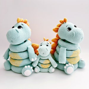May include: Three crocheted plush toys shaped like dragons. The largest dragon is light blue with yellow accents and has a curled tail. The middle dragon is light blue with yellow accents and has a straight tail. The smallest dragon is light blue with yellow accents and has a straight tail.