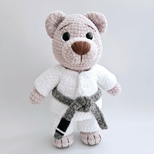 Może przedstawiać: Pluszowy miś szydełkowy w białym stroju karate z czarnym paskiem. Miś ma brązowe oczy i brązowy nos.