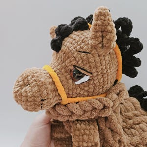 Pu&ograve; includere: Un giocattolo a forma di cavallo fatto a mano, in morbido peluche marrone, con criniera e coda nere. Il cavallo ha un'imbracatura gialla e un occhio dettagliato. Il materiale morbido e strutturato gli conferisce un aspetto coccoloso.