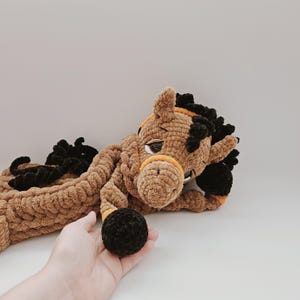 Handmade crochet horse pajama holder in brown plush yarn, black hooves, orange bridle, shown held in a human hand for scale