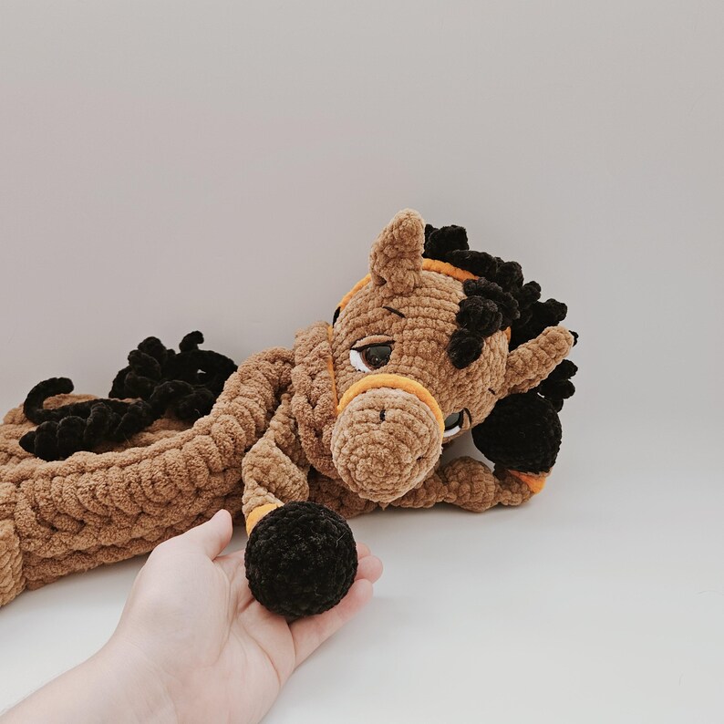 Handmade crochet horse pajama holder in brown plush yarn, black hooves, orange bridle, shown held in a human hand for scale