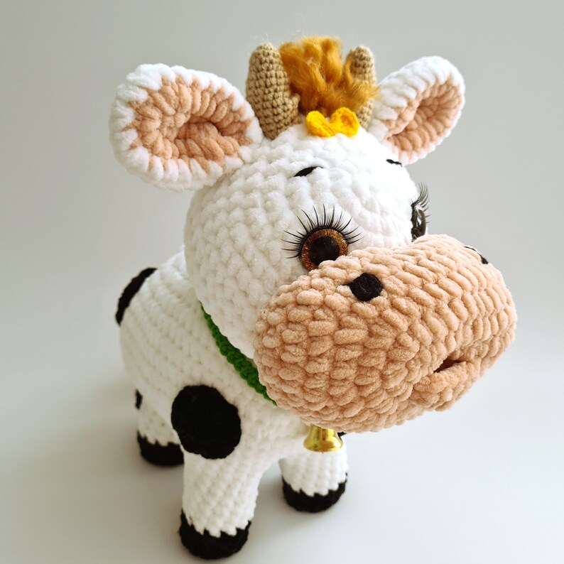 May include: White crocheted cow plush toy with black spots, brown fuzzy nose, and green collar. The cow has a yellow flower on its head and brown eyes with long eyelashes.