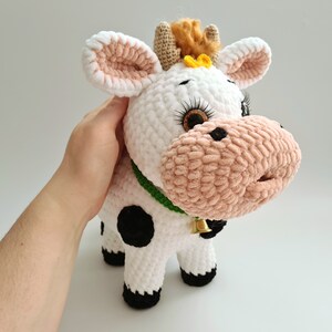 May include: A white crocheted cow plush toy with black spots, pink ears, and a green collar with a gold bell. The cow has brown eyes with eyelashes and a yellow flower on its head.