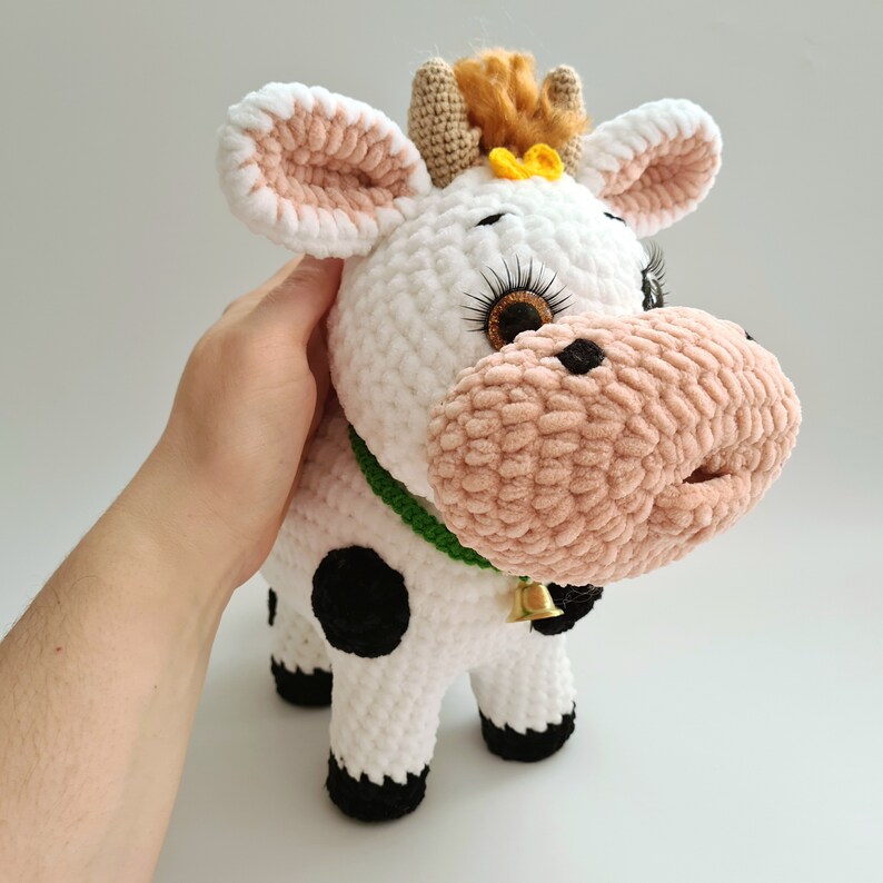 May include: A white crocheted cow plush toy with black spots, pink ears, and a green collar with a gold bell. The cow has brown eyes with eyelashes and a yellow flower on its head.