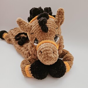 Crochet brown horse pajama bag plush toy, lying down with front arms under head, looking straigh ahead, black mane and orange bridle details