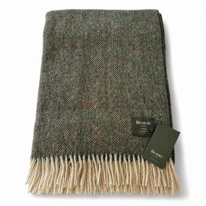 May include: A folded green and brown woven throw blanket with a cream-colored fringe. The blanket has a textured appearance and includes a tag that reads "Bronte by Moon."