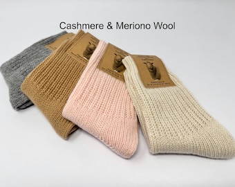May include: Four pairs of ribbed knit socks in various colours: grey, tan, pink, and cream. Each pair has a tag that reads "Cashmere" and "Merino Wool". The socks are made of a soft, warm material.