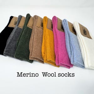 May include: A collection of eight pairs of ribbed Merino wool socks arranged in a row, featuring colours including black, charcoal, dark green, tan, mustard yellow, magenta, light blue, and white, each with a brown paper label attached.