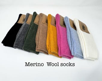 May include: A collection of eight pairs of ribbed Merino wool socks arranged in a row, featuring colors including black, charcoal, dark green, tan, mustard yellow, magenta, light blue, and white, each with a brown paper label attached.