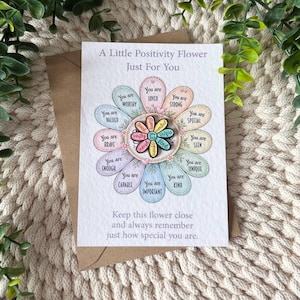 May include: A white greeting card featuring a printed illustration of a flower with pastel-coloured petals, each containing positive affirmations like You are loved and You are strong. A small metal flower pin is attached to the centre.