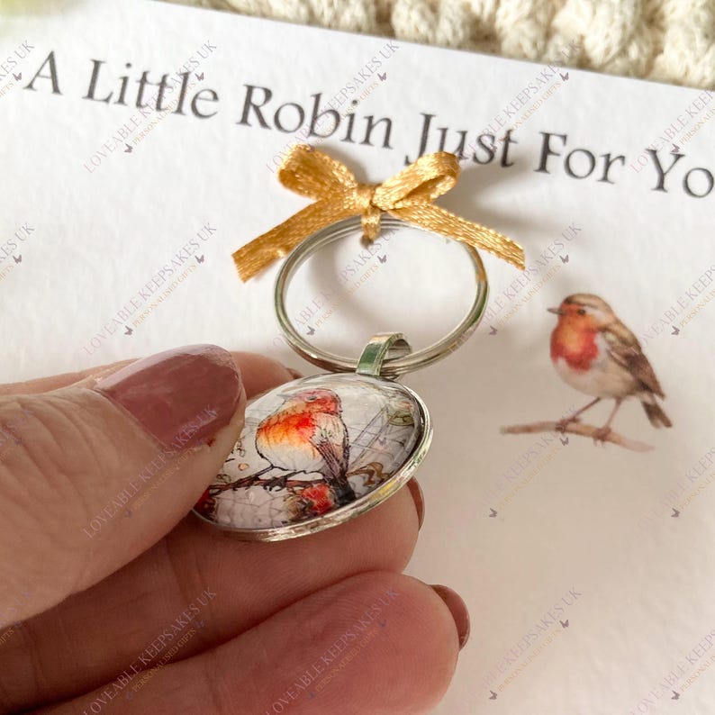 Puede incluir: Un llavero plateado con un colgante ovalado que muestra una ilustraci&oacute;n de acuarela de un petirrojo. Se adjunta una cinta dorada. El texto "A Little Robin Just For You" es visible.