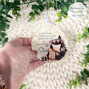 May include: A circular Christmas decoration with a printed design of a comfortable armchair, a Christmas tree, and a gift. The ornament reads "An empty chair, but a heart full of love, missing you this Christmas Steve." A white ribbon is attached.