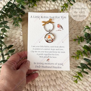A keyring with a robin bird pendant, presented on a card with the message "A Little Robin Just For You". The card includes a poem and is set against a cream-colored woven background. The text reads "In loving memory of Your Dear Husband.