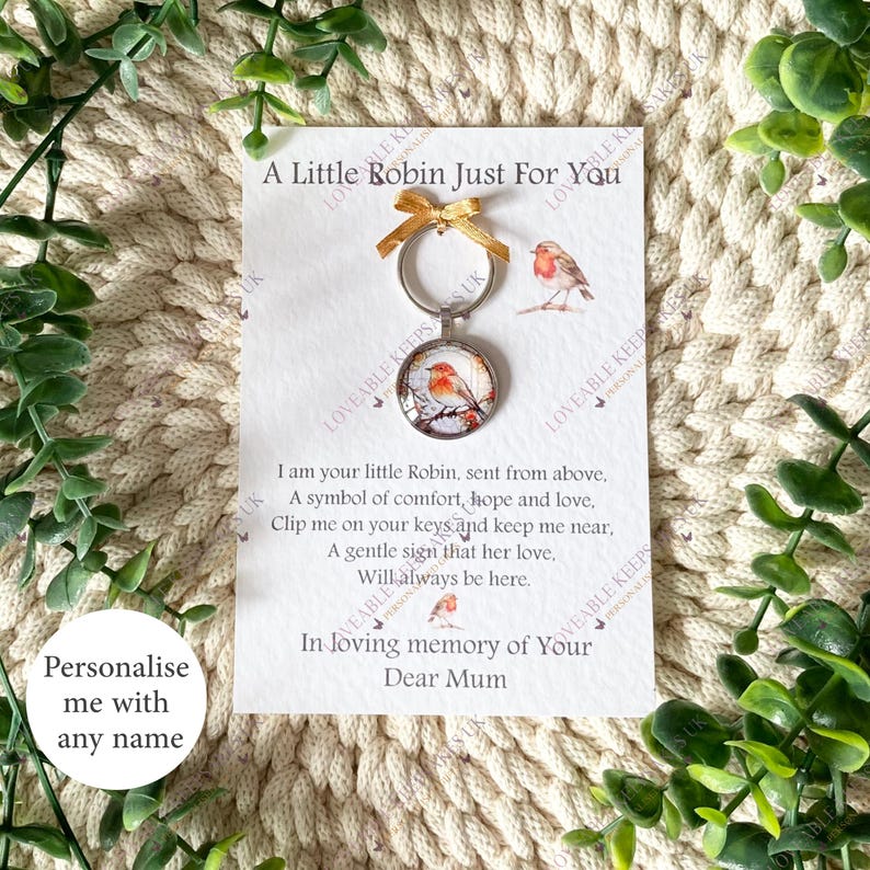 Puede incluir: Un llavero con un colgante redondo con una ilustraci&oacute;n de un petirrojo. El llavero est&aacute; adjunto a una tarjeta con el texto "A Little Robin Just For You" y un mensaje de recuerdo. El fondo es una textura tejida de color crema. El texto dice "In loving memory of Your Dear Mum".