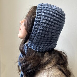 May include: A blue knitted hood and scarf set. The hood is a square shape with a ribbed texture, and the scarf hangs down the back. The set is made of a soft, textured yarn.