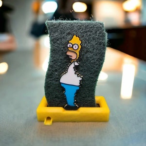 May include: A yellow cartoon character with a blue shirt and blue trousers is attached to a grey sponge. The sponge is in a yellow plastic holder.