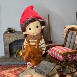 May include: A handmade doll dressed in a red felt hat, brown overalls, and a striped shirt. The doll has a scarf, rosy cheeks, and freckles. The doll is standing in front of a fireplace and a wooden chair with a plaid pillow.