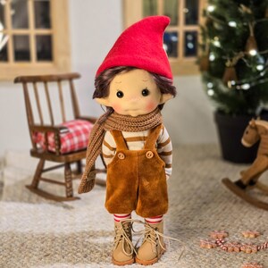 May include: A handmade doll dressed in a red pointed hat, brown scarf, and corduroy overalls. The doll has brown hair, rosy cheeks, and is wearing striped socks and tan boots. A wooden rocking chair and Christmas tree are in the background.