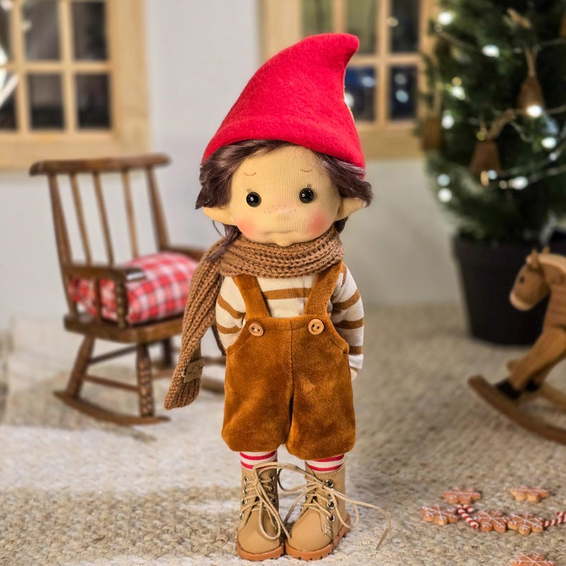 May include: A handmade doll dressed in a red pointed hat, brown scarf, and corduroy overalls. The doll has brown hair, rosy cheeks, and is wearing striped socks and tan boots. A wooden rocking chair and Christmas tree are in the background.