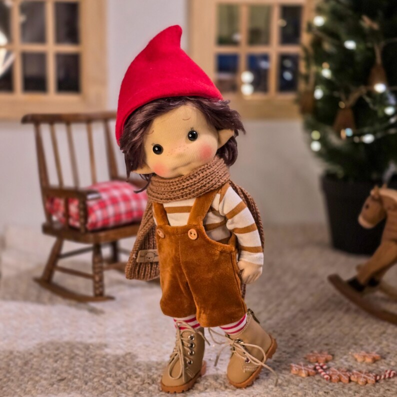 May include: A handmade doll dressed in a red pointed hat, brown overalls, and a striped shirt. The doll has brown hair, a knitted scarf, and tan boots. The doll is standing on a light-colored rug, with a rocking chair and Christmas tree in the background.