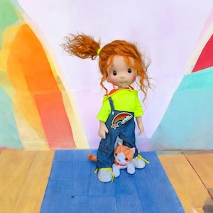 May include: A handmade doll with red hair in a ponytail, wearing a neon green shirt, denim overalls with a rainbow patch, and white shoes. A small stuffed orange and white cat is at the doll's feet. The background has colorful painted shapes.