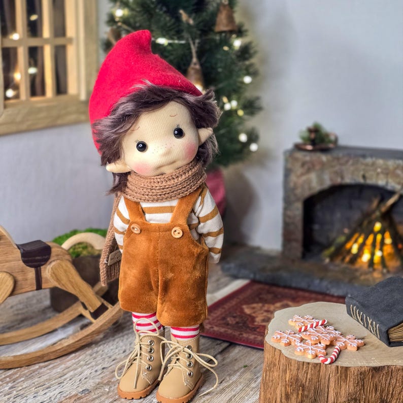May include: A handmade doll dressed in a red elf hat, brown hair, and a tan scarf. The doll wears a striped shirt, brown overalls, and tan boots. The background includes a Christmas tree, a fireplace, and gingerbread cookies, creating a festive holiday scene.